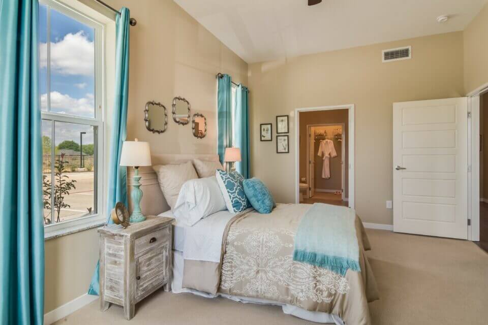 Model apartment bedroom with beige and teal bedding and two windows on either side of bed, teal curtains