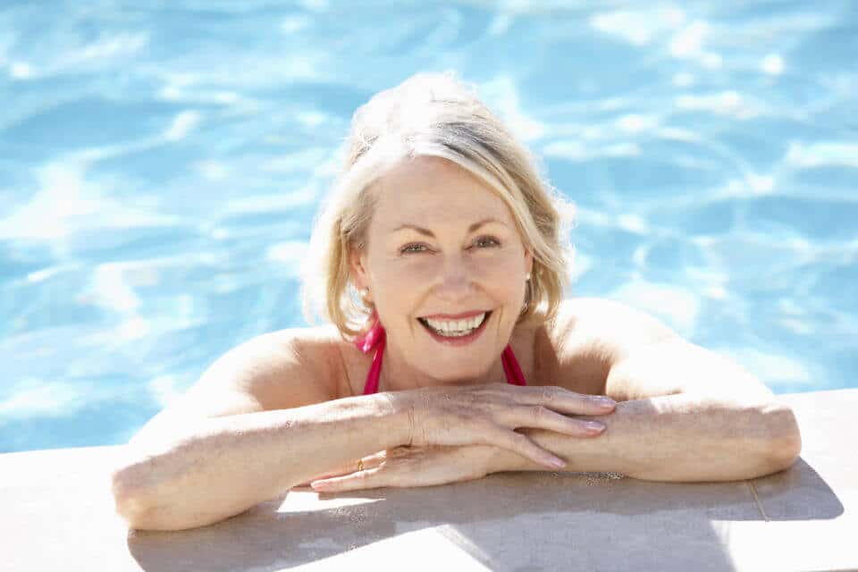 Senior woman in outdoor pool smiling with arms crossed on edge of pool.